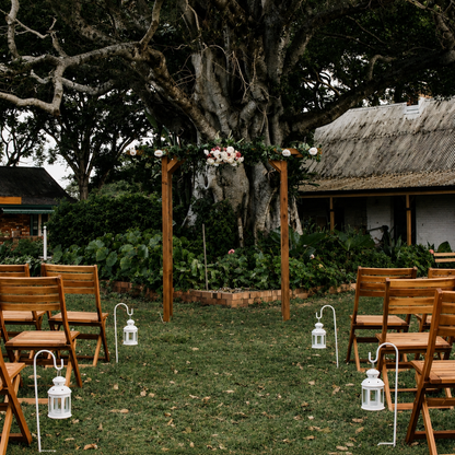 Outdoor wedding setup with wooden chairs, floral arch, and lanterns under a large tree.